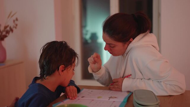 Mother points to school worksheet while guiding young son at dining table in family home, encouraging focused learning, patience, confidence, knowledge and parental support