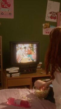 Vertical shot of Caucasian teenage girl seen from behind sitting on bed, eating popcorn and watching retro show on CRT television. Ideal for nostalgia marketing and retro lifestyle campaigns