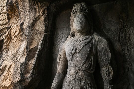 An ancient unfinished stone statue of a deity with a worn face in a rocky alcove.