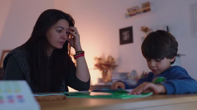 Mother supervising schoolboy reviewing an activity workbook at home dining table, supporting education, patient guidance, concentration and parent-child connection
