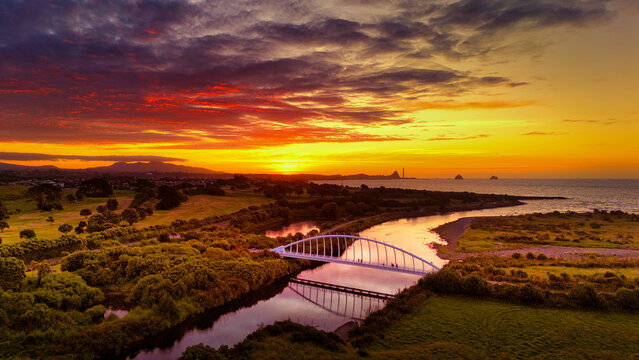 Aerial view of Te Rewa Rewa Bridge spanning the river at sunset with vibrant orange clouds reflecting in the water and coastal landscape in New Plymouth, Taranaki Region, New Zealand.