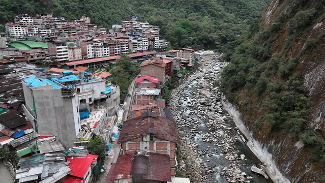 Aerial view of the town of Aguas Calientes nestled in a deep valley beside a rocky river with dense buildings and steep mountain slopes in Aguas Calientes, Cuzco, Peru.