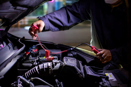 Selective focus to battery positive terminal and mechanic connecting red booster jumper cables to a car battery under the open hood at night. Concept of roadside assistance and auto repair service.