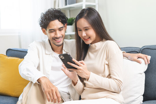 Happy couple love at home, two asian young spending time, bonding to each other romantic on sofa in living room while man embrace woman using smartphone, mobile phone to selfie take a photo together.