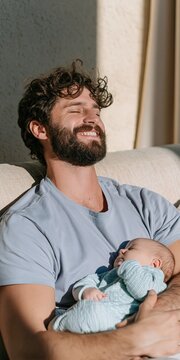 Smiling father holding a sleeping baby on a couch in sunlight for parenting background and family wellness design