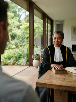 Judge in black robes at a table before windows onto a lush garden in soft daylight
