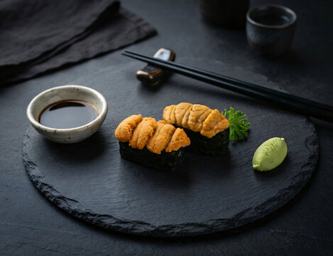 Decadent bafun uni gunkan sushi duo featuring vibrant orange sea urchin roe on crisp nori served on black slate with shoyu, wasabi and chopsticks.