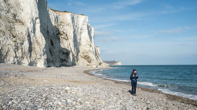 Person stands on a pebble beach observing the sea beneath tall white chalk cliffs