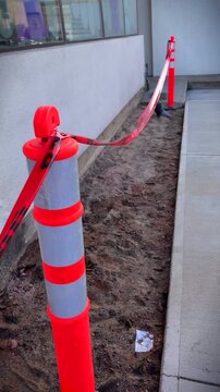 Red, tall, safety markers with caution tape cordoning off unpaved area next to an exterior wall. In vertical format.