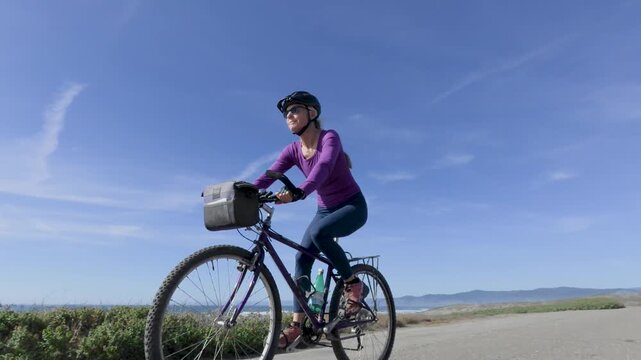 Mature woman rides her bike along a trail next to the ocean in Fort Bragg, California on a sunny day enjoying the fresh air.