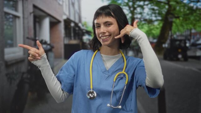 Woman doctor in blue scrubs with yellow stethoscope holds hand to ear for call gesture on city street; compassion.