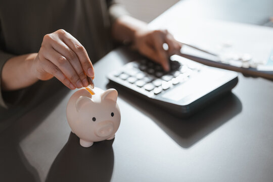 A hand drops a coin into a ceramic bank beside a calculator as a budget planner records deposits and tracks savings toward future household needs and planned purchases through regular contributions.