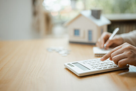 A person uses a calculator beside a model house while writing figures to estimate mortgage costs and compare each loan option before choosing an affordable monthly amount for a planned purchase.