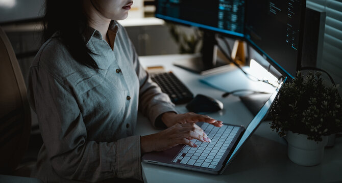 A woman types code on a laptop at a multi-monitor desk while a data upload runs in the background and a progress indicator tracks work saved to a remote server during an evening programming session.
