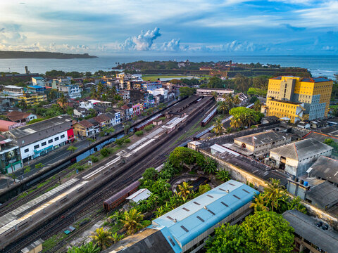 Aerial view of Galle Railway Station with trains on tracks, surrounded by urban buildings, lush greenery, and the coastal landscape of Galle, Southern Province, Sri Lanka.