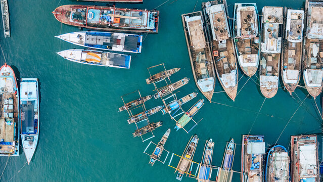 Aerial view of numerous fishing boats moored in the turquoise harbor waters, showcasing traditional outrigger designs and wooden hulls Palabuhanratu, West Java, Indonesia.