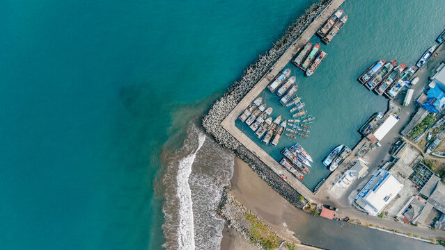 Aerial view of a harbor with numerous fishing boats moored at the pier, turquoise water, and a breakwater along the coast in Palabuhanratu, West Java, Indonesia.