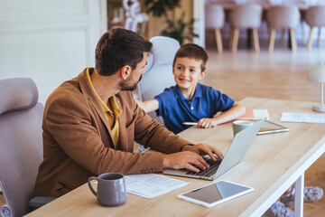 Fototapeta premium Father Working From Home With Son At Home Office Desk Sharing A Moment While He Works
