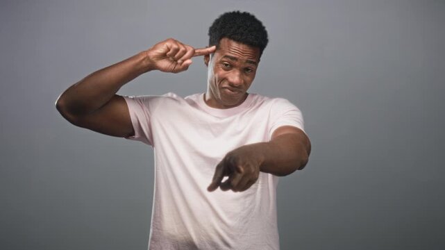 Man pointing finger to temple and pointing forward while smirking, wearing pale pink t shirt against gray studio backdrop; playful confidence.