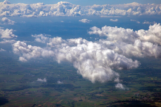 An aerial view of a cloudscape,  scattered puffy cumulus or stratocumulus clouds hovering above a patchy, green landscape visible on the ground below. Mid-to-low level detached cumulus clouds. 