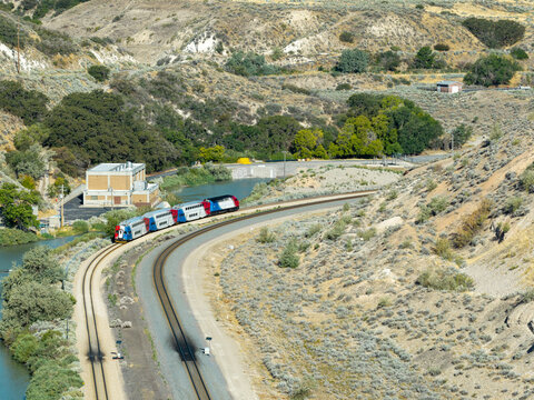 Aerial view of a commuter train traveling along the tracks beside the Jordan River in a rugged canyon landscape in Bluffdale, Utah, United States.
