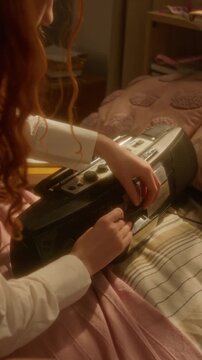 Vertical video of young Caucasian woman inserting cassette tape into vintage boombox on bed. Footage is ideal for retro lifestyle ads, nostalgia marketing, and music history projects