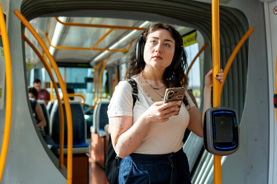 Woman standing in a tram, holding smartphone, wearing headphones, commuting in the city