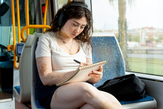 Young woman with vitiligo riding tram, listening music, writing in notebook