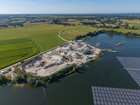Aerial view of a sand and gravel extraction site with large piles of aggregate and machinery situated next to a lake with a floating solar array in the Netherlands.