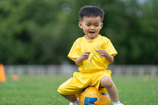 Joyful Asian child sitting on football during daytime sports practice on green grass field, authentic playful toddler activity