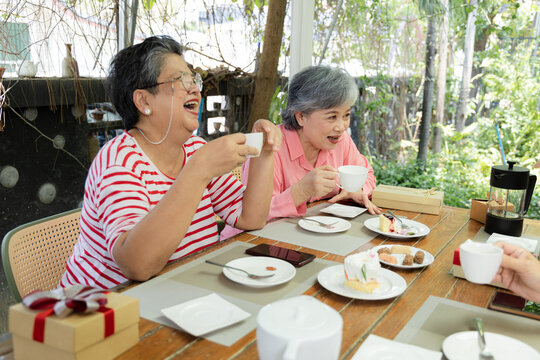 Senior women laugh over hot tea during outdoor cafe reunion, sharing desserts and conversation beside wrapped gifts as friendship and relaxed companionship fill cheerful gathering, aging society.