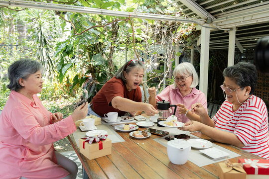 Elderly friends enjoy sharing cake at garden cafe reunion, reaching across table with forks and plates while laughter, tea, and wrapped gifts create joyful social bonding outdoors, aging society.