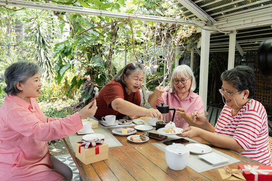 Senior group shares dessert at outdoor cafe reunion, passing cake across table while laughing together among tea cups, gift boxes in warm celebration of friendship and companionship, aging society.