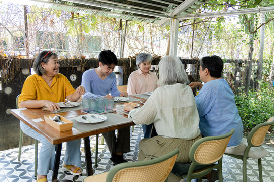 Senior women and young man gather at outdoor cafe for reunion, sharing snacks, conversation, and laughter while friendship and social connection create warm intergenerational time, aging society.