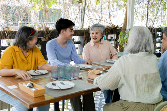 Active seniors enjoy outdoor cafe reunion with young man, smiling and talking around table while snacks and conversation strengthen friendship, community, intergenerational connection, aging society.