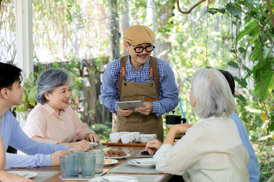 Senior group listens as waiter uses tablet during outdoor cafe reunion, combining attentive restaurant service, conversation, refreshments with relaxed friendship around garden table, aging society.