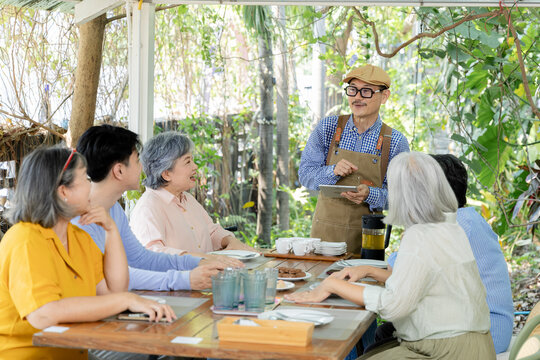 Retired seniors meet waiter using tablet at garden cafe, discussing service and refreshments around table while friendly conversation supports relaxed reunion and social connection, aging society.
