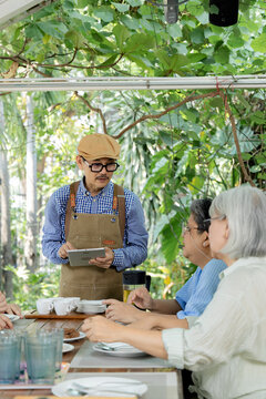 Mature women speak with waiter holding tablet at garden cafe, discussing refreshments during reunion while friendly service, conversation create comfortable social connection outdoors, aging society.