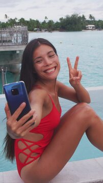 Social media travel influencer taking selfies at luxury hotel infinity pool overwater bungalow in fashion swimsuit. Asian beauty woman on vacation. Raw vertical video of real woman without makeup