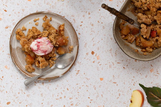 Apple crumble with a scoop of ice cream on a plate and a baking dish on a terrazzo table
