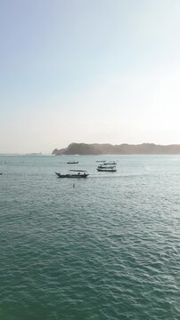 Aerial drone approaching coastal headland with surf break and wooden boats in turquoise bay Kuta Lombok