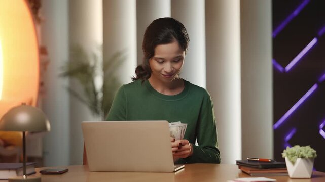 Pleased young Korean woman counts money by laptop, celebrating business profit
