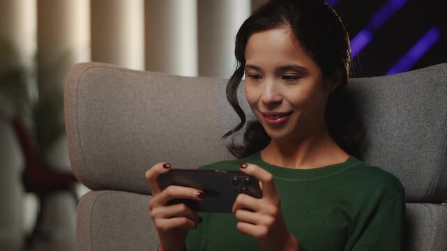 Happy young Asian woman plays online game on cellphone, relaxing at home