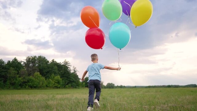 Child running with colorful balloons across green field. Happy boy holds bunch of bright balloons in summer meadow. Kid runs freely under cloudy sky. Balloons float as child plays outdoors in nature.