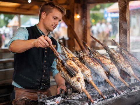 Koch bestreicht ganze Fische am Holzspie&szlig; &uuml;ber gl&uuml;hender Holzkohle. Traditionelle Zubereitung von Steckerlfisch am Volksfest mit Rauch, Grillhitze und rustikalem Imbissstand.