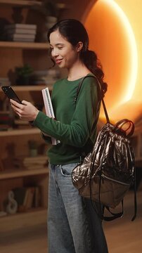 Cheerful Asian female student scrolls social media on phone in modern library