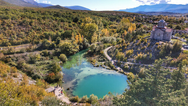 Aerial view of the Eye of the Earth spring surrounded by lush trees and a historic church on a hill with people walking nearby in Cetina, Sibenik-Knin County, Croatia.