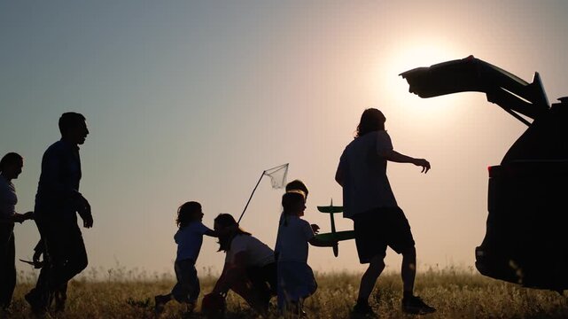 Happy family plays in field at sunset near open car trunk. Silhouettes of children and parents run and enjoy outdoor activity. Family plays ball in meadow at golden sunset. Kids run together.