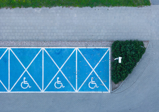 Aerial view of blue painted handicap parking spaces with white markings and wheelchair symbols next to a green shrubbery area in Gdansk, Pomeranian Voivodeship, Poland.