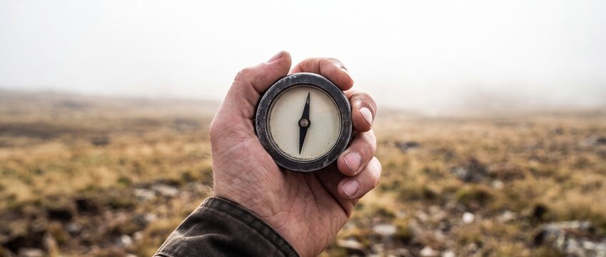 A male hand holds old metal compass with an arrow pointing forward, surrounded by dry grass field and foggy sky during daytime.